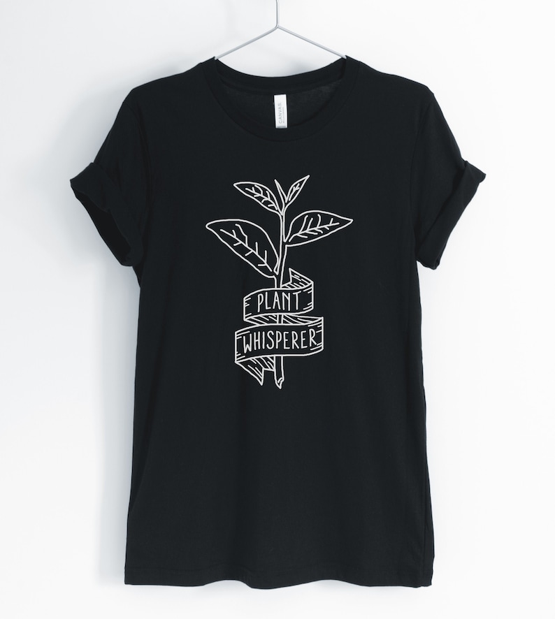 Puede incluir: Camiseta negra con un gr&aacute;fico blanco de una planta con una pancarta que dice "Plant Whisperer".