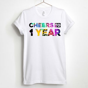 May include: White t-shirt with the text "CHEERS TO 1 YEAR" in colourful letters with a rainbow splatter effect.