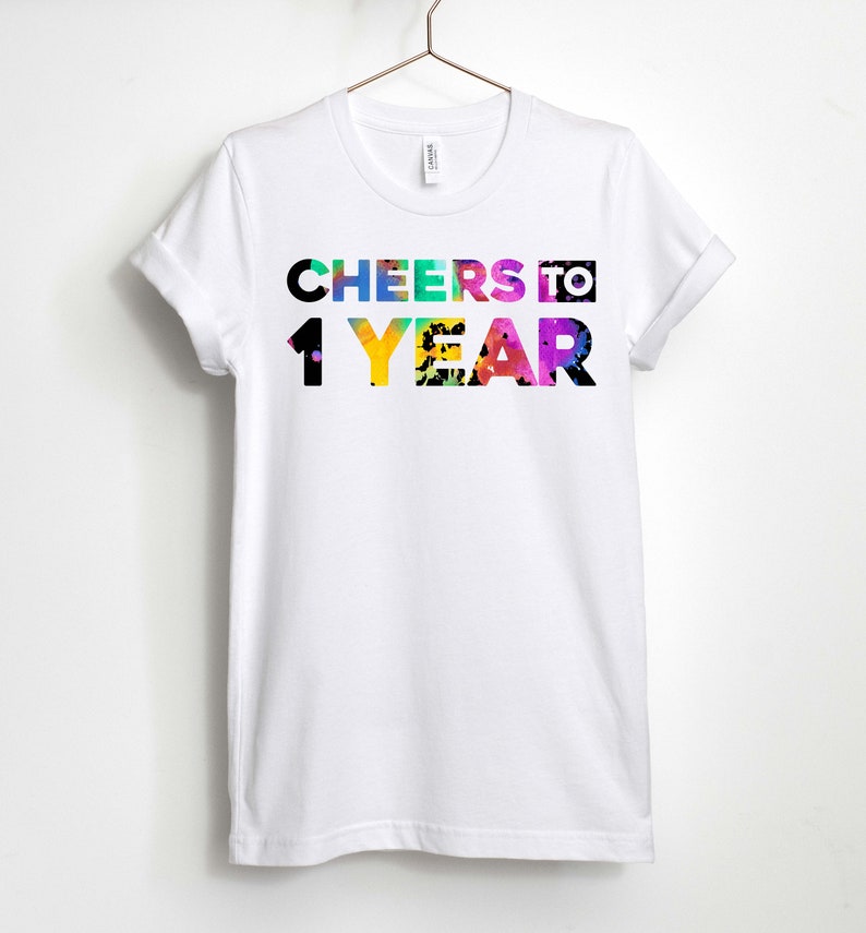 May include: White t-shirt with the text "CHEERS TO 1 YEAR" in colourful letters with a rainbow splatter effect.