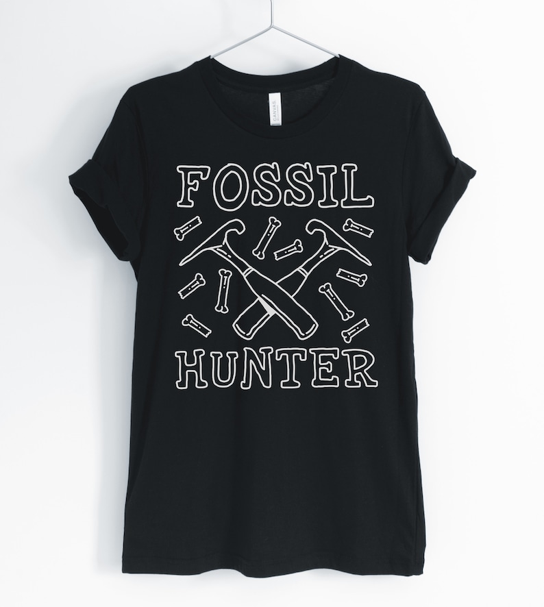 May include: Black t-shirt with white text that reads "Fossil Hunter". The design includes two crossed pickaxes and small bone shapes.