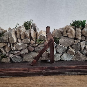 May include: A miniature stone wall model constructed from small, irregular gray and tan rocks, featuring a dark brown wooden stile integrated into the center and small patches of green moss tucked between the stones, all mounted on a rectangular wooden base.