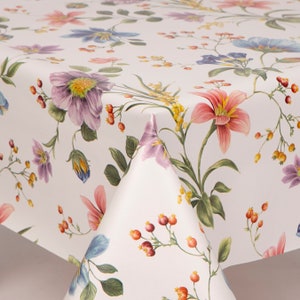 May include: A white tablecloth with a vibrant floral pattern. The design features a variety of flowers in shades of pink, purple, blue, and yellow, along with green leaves and red berries. The tablecloth is folded, showcasing the detailed print.