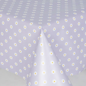 May include: A lilac tablecloth featuring a repeating pattern of white daisies with yellow centres. The tablecloth is folded, revealing the pattern on the surface and the underside.