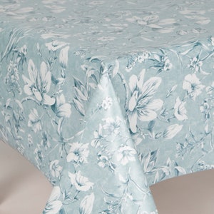 May include: A pale blue tablecloth with a floral design. The pattern includes large white flowers and smaller white blossoms with green and blue leaves. The tablecloth is folded, revealing the repeating pattern.