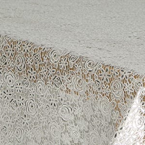 May include: A white lace tablecloth with a floral pattern, featuring roses and other flower designs. The intricate openwork creates a delicate and elegant texture. The tablecloth is draped over a wooden surface.