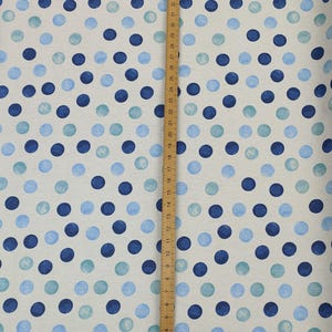 May include: A fabric sample featuring a pattern of blue polka dots in shades of navy, light blue, and teal, set against a neutral backdrop. A wooden ruler with centimetre markings is placed vertically on the fabric.