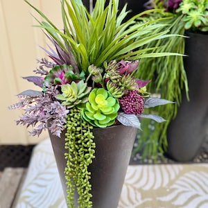 May include: A tall, dark gray planter filled with a variety of artificial succulents and greenery. The arrangement includes bright green, purple, and gray-green plants, with long, trailing green vines. A second planter is visible in the background.
