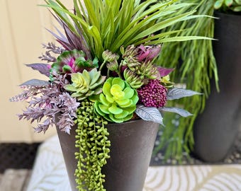 May include: A tall, dark gray, tapered conical planter filled with an arrangement of artificial plants. The display features bright green spiky grass, various succulent rosettes in shades of lime and deep purple, and trailing light green vines.