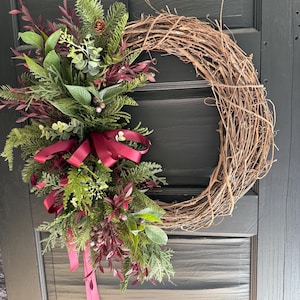 Peut inclure: Une couronne circulaire faite de brindilles brunes naturelles, ornée d'un arrangement asymétrique d'aiguilles de pin vertes, de feuilles d'eucalyptus et de feuillage bordeaux foncé. Un ruban de satin rouge foncé forme un nœud aux extrémités tombantes.