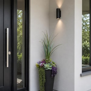 May include: A black outdoor planter filled with greenery and purple flowers stands beside a black door. A black wall-mounted light fixture is above the planter. The door has a long silver handle.