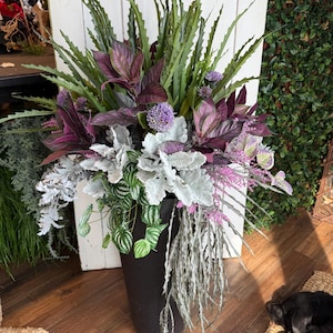 May include: A tall black cylindrical floor vase filled with an arrangement of artificial plants, including spiky green leaves, deep purple foliage, silver-grey dusty miller leaves, small purple globe flowers, and trailing thin grey-green stems.