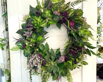 May include: A lush green and purple floral wreath with succulents and ferns. The wreath is hanging on a white door.