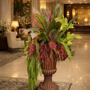 May include: A large, brown, fluted pedestal urn filled with a lush arrangement of greenery. The display features broad, pink-tipped leaves, bright green fern fronds, and cascading, textured, deep red foliage that hangs over the urn's rim.