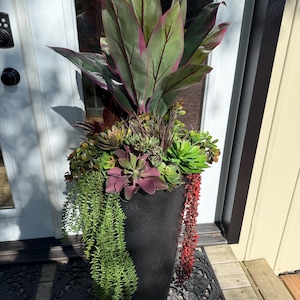 May include: A tall, black, cylindrical planter filled with an arrangement of artificial plants, including large, pointed green and magenta leaves, various green and purple succulents, and trailing green and red foliage cascading down the sides.