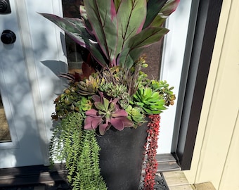 May include: A tall, black, cylindrical planter filled with an arrangement of artificial plants, including large, pointed green and magenta leaves, various green and purple succulents, and trailing green and red foliage cascading down the sides.