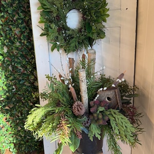 May include: A decorative arrangement with a green wreath and a white centre, and a potted display with faux greenery, birch logs, a birdhouse, and pinecones. The arrangement is set on a wooden stand, creating a natural, woodland theme.