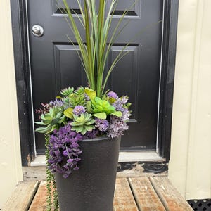 May include: A tall, black, textured planter filled with artificial plants. The arrangement includes green succulents, purple cascading flowers, and tall green grass-like stems. The planter sits on a weathered wooden surface.