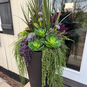 May include: A tall, dark brown planter filled with artificial plants. The arrangement includes large green succulents, long green grass-like blades, purple flowers, and trailing green vines. The planter is placed on a wooden surface.