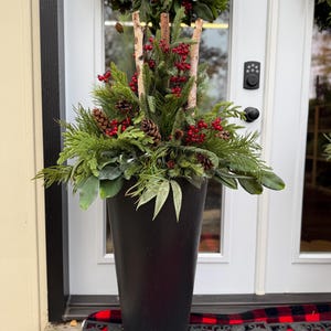 May include: A tall, black planter filled with evergreen branches, red berries, and pinecones. Birch branches are used as a decorative element. A matching wreath hangs above the door. The arrangement is festive and suitable for the holiday season.