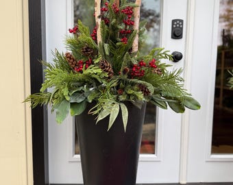 May include: A tall, matte black tapered planter filled with an arrangement of evergreen boughs, pinecones, bright red berries, and three vertical birch wood branches. The arrangement is dense and textured, featuring various shades of green foliage.