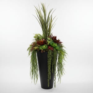 May include: A tall, matte black tapered planter filled with an arrangement of artificial greenery. The display features upright spiky grass, various green and deep red succulents, and long, trailing vines of small, bead-like foliage cascading down.