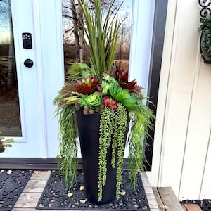 May include: A black, cylindrical planter overflowing with artificial plants. The arrangement features tall, green grass-like foliage, red and green succulents, and trailing green vines. The planter sits on a decorative black mat on a wooden porch.