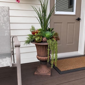 May include: A brown pedestal planter with a fluted base and rim, filled with an arrangement of artificial green succulents, tall spiky grass-like foliage, and a trailing string of pearls plant, creating a dense and varied botanical display.