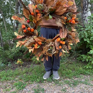 Peut inclure: Une grande couronne circulaire composée de feuillage d'automne séché, avec des feuilles de magnolia brunes, des cosses de lanterne chinoise orange, des panaches plumeux brun foncé et de petites fleurs séchées disposées en un arrangement dense.