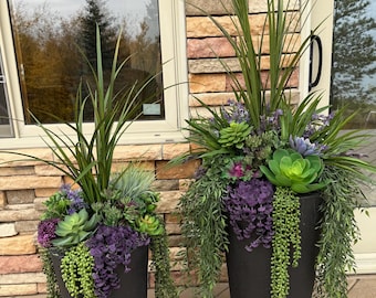 May include: Two tall, matte black cylindrical planters filled with an arrangement of artificial greenery. The plants include long, spiky green grass, various green and purple succulents, and trailing strings of small green beads.