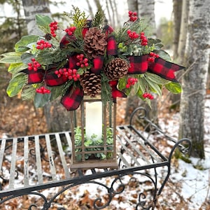 Puede incluir: Una linterna decorativa con una vela encendida, adornada con piñas, bayas rojas y vegetación, acentuada por una cinta a cuadros roja y negra. La linterna se encuentra sobre un banco de metal negro con detalles en espiral.
