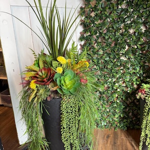 May include: A black, cylindrical planter overflowing with artificial plants. The arrangement includes succulents, grasses, and trailing greenery in shades of green, yellow, and red. The planter is placed on a decorative, dark metal base, creating a natural look.