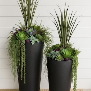 May include: Two tall, matte black tapered planters filled with an arrangement of artificial greenery. The plants include upright spiky grass, various green and purple succulents, and trailing vines that drape over the sides of the containers.
