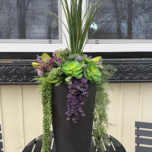 May include: A black, textured planter filled with artificial plants. The arrangement includes green succulents, purple hanging flowers, and tall green grass-like foliage. The planter sits on a black metal table. The planter is approximately 60 cm tall.