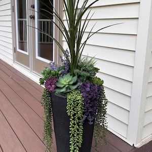 May include: A tall, black, tapered cylindrical planter filled with an arrangement of artificial plants, including long green grass blades, various green succulents, purple flowering sprigs, and trailing green string-of-pearls vines.