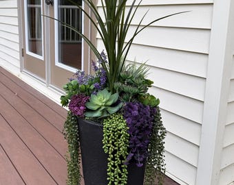 May include: A tall, black, tapered cylindrical planter filled with an arrangement of artificial greenery. The display features long, spiky green leaves, various green succulents, purple floral clusters, and trailing green string-of-pearls vines.