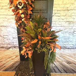 May include: A tall black tapered planter filled with a lush arrangement of green and orange foliage, including croton leaves, ferns, and a central bromeliad, paired with a matching vertical door swag featuring orange ribbons and white pumpkins.