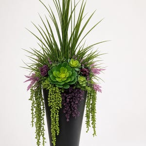 Peut inclure: Un grand pot conique noir mat rempli d'un arrangement de verdure artificielle. La composition comprend de longues herbes vertes pointues, des rosettes succulentes vert vif et des tiges tombantes de petit feuillage vert et violet foncé.