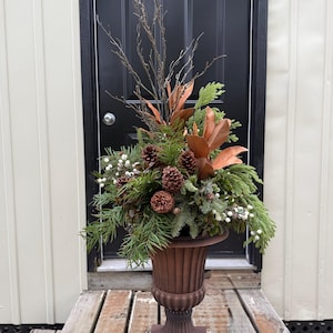 May include: A decorative floral arrangement in a dark brown fluted pedestal urn. The arrangement features green pine branches, brown pinecones, copper-coloured magnolia leaves, bare twigs, and clusters of small white berries, creating a full display.