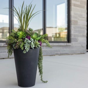 Peut inclure: Un grand pot noir rempli de plantes et de succulentes artificielles. L'arrangement comprend des feuillages verts, violets et gris, avec de longues feuilles vertes et fines au centre. Le pot est posé sur une surface en béton.