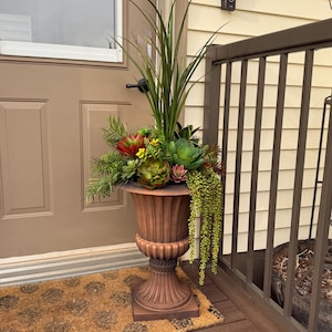 May include: A large, ornate brown urn planter filled with a variety of faux succulents and greenery. The planter is placed on a brown welcome mat and a wooden deck, near a door and railing.