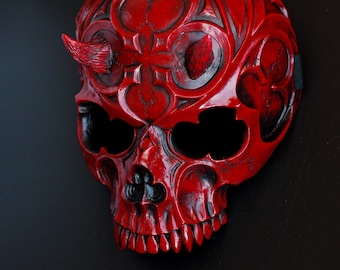 May include: A red skull mask with intricate carvings and a pair of horns. The mask is made of a hard material and has a glossy finish.