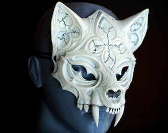 Könnte beinhalten: Eine weiße, detaillierte und komplizierte Maske mit katzenartigem Design. Die Maske hat scharfe Zähne und ein geometrisches Muster.