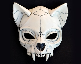 Könnte beinhalten: Weiße Katzenmaske mit schwarzen Details, die einem Schädel mit Zähnen ähnelt. Die Maske hat Augenlöcher und eine kleine schwarze Nase.