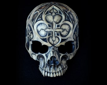 May include: A white skull mask with a black and white geometric design. The mask has a detailed design of a cross and hearts.