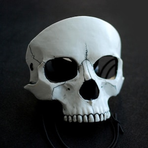 Op de afbeelding: Een wit schedelmasker met zwarte oogkassen, neusholte en tanden. Het masker heeft zichtbare scheuren en een zwart touwtje. Het masker staat tegen een donkere achtergrond.