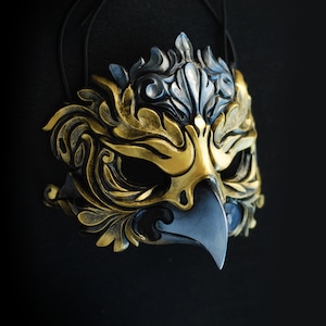 May include: A decorative mask in the shape of a bird's head, featuring gold, black, and silver tones. The mask has intricate, ornate details and a beak-like design. It is suspended against a black background, suggesting a theatrical or costume theme.