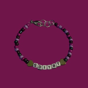 May include: A beaded bracelet with a mix of purple, dark pink, and black beads. The bracelet features white letter beads spelling "SDIYBT" and two green heart-shaped charms. The clasp is silver.