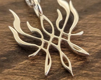 May include: A silver pendant necklace featuring an openwork design. The pendant has a symmetrical, stylized flame-like shape with a central geometric pattern. The necklace chain is also silver and appears delicate. The background is a dark wood surface.
