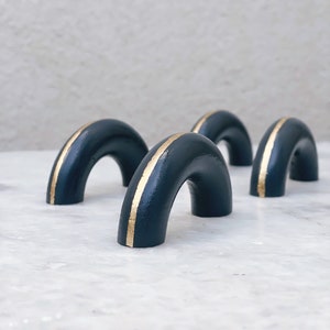 Könnte beinhalten: Drei schwarze Keramik-Schubladengriffe mit einem goldenen Streifen in der Mitte. Die Griffe sind bogenförmig.