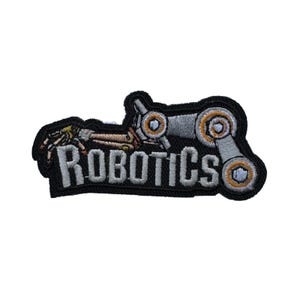 May include: Embroidered patch with the word "ROBOTICS" in white lettering. The patch features a stylized robotic arm and other mechanical elements in shades of gray, gold, and black. The patch has a black border.